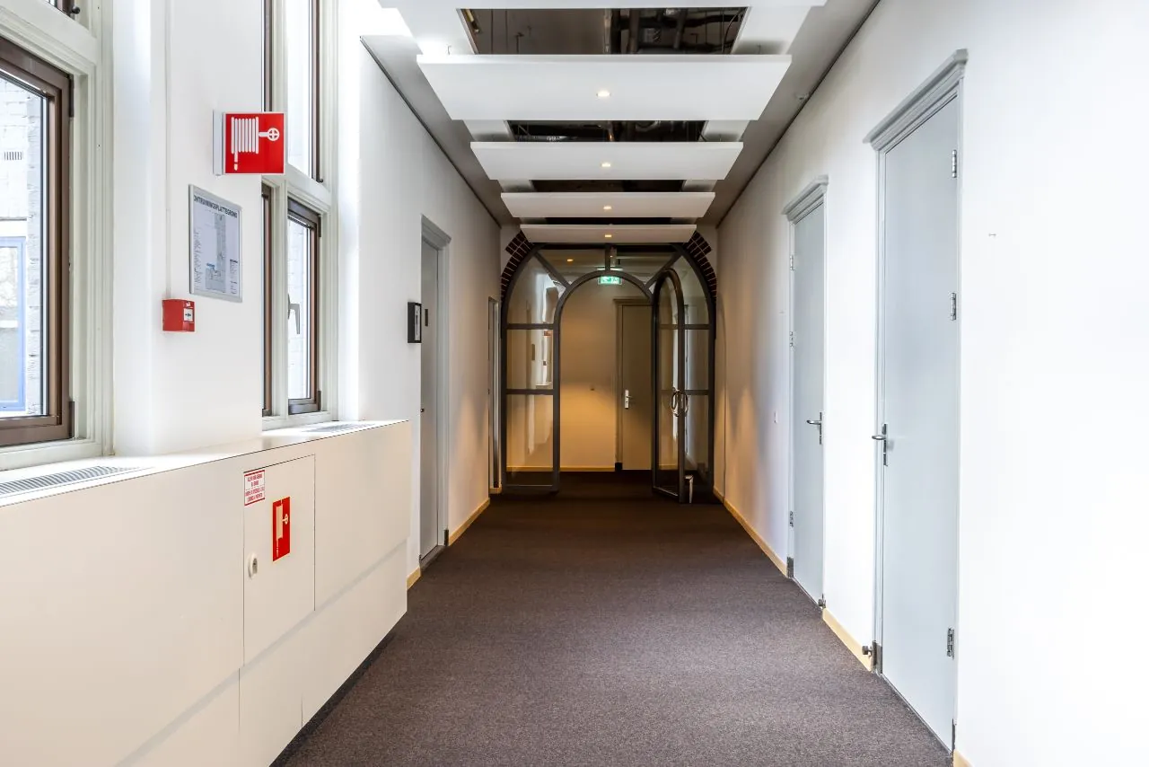 Geverfd wit kantoorinterieur met grijze deuren en glazen boogdeur aan het eind van de gang op de Lage Naarderweg.